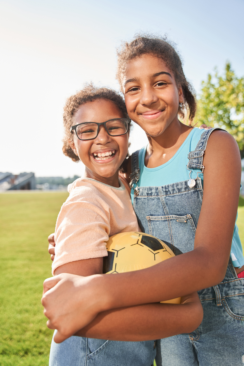 Little black girlfriends standing with football ball and hugging outdoors. Concept of childhood. Idea of friendship. Modern kid lifestyle. Joyful curly brunette girls looking at camera. Sunny daytime