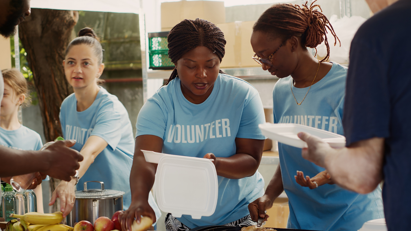 At outdoor food bank, multiethnic female volunteers provide free food to homeless men. Voluntary ladies provide humanitarian relief and create community by assisting the hungry and underprivileged.
